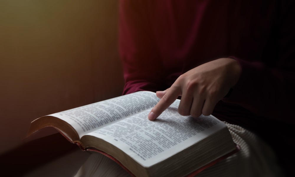 The hand of a person studying a Bible open on their lap, tracing their finger along the text