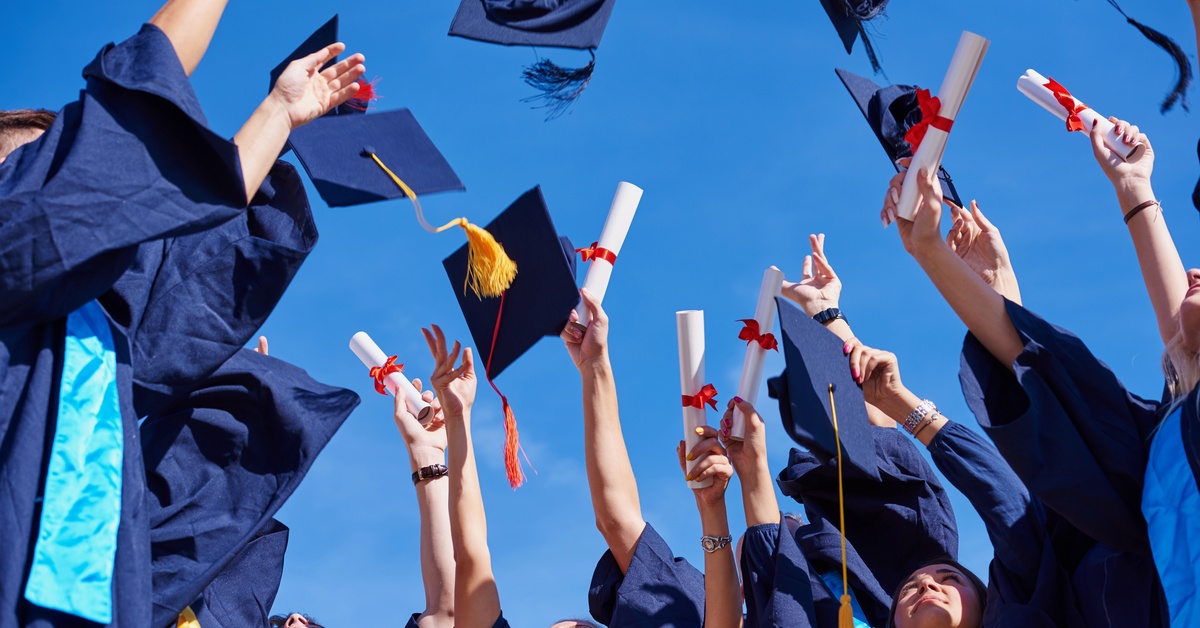 Graduates in navy blue gowns raising their arms in celebration. Some throw their caps up while others raise their diplomas.