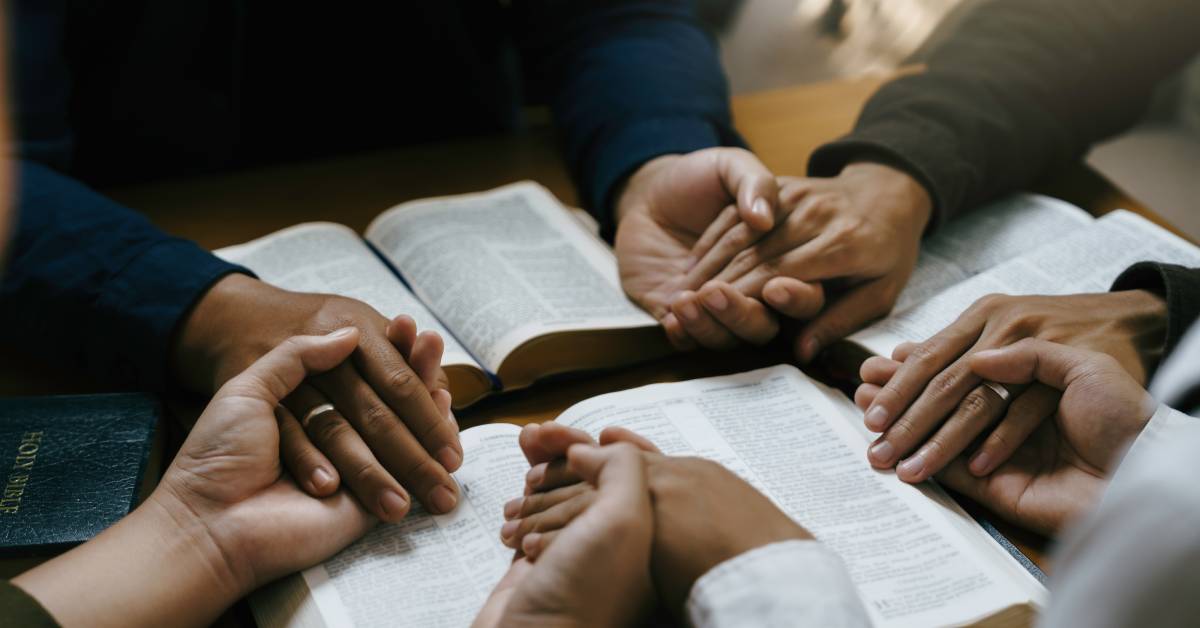 People seated around a table hold each other's hands in prayer. There are three open Bibles on the table and one closed one.