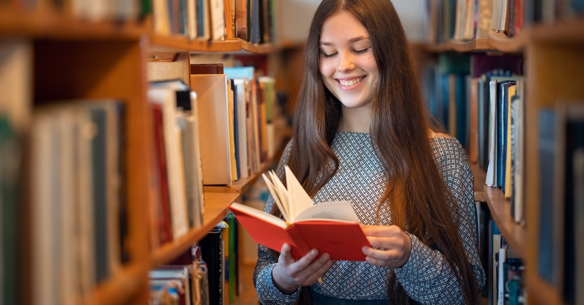 A young woman stands between long rows of full bookshelves. She's holding a book and smiling as she looks at a page.