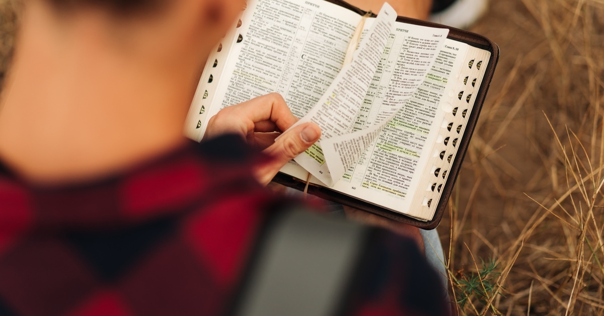 Overhead view of a person sitting outdoors, turning the pages of a Bible. The text is printed in two columns per page.