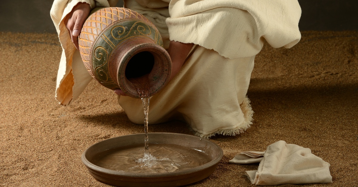 A person wearing a robe pours water from a clay jug into a shallow basin on the ground. A clean cloth is next to the basin.