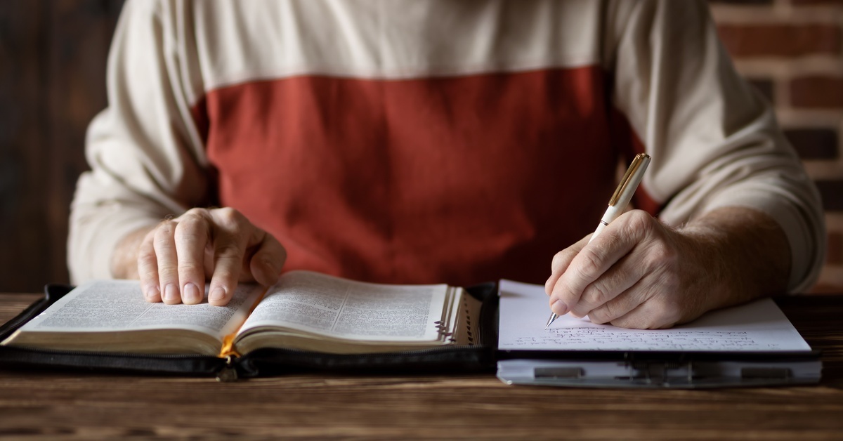 A person sitting at a table, with their fingers tracking a Bible verse. With their other hand, they write notes on a notepad.
