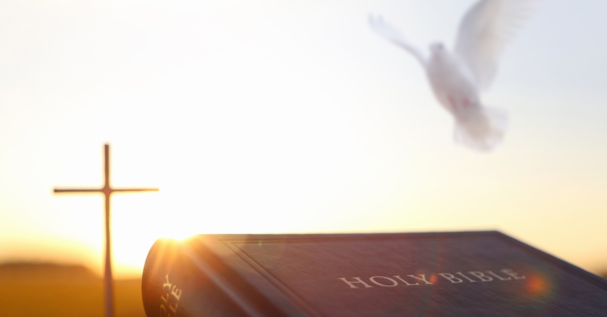 A Holy Bible lies in the foreground, and a white dove flies above. In the background, the sun sets behind a cross.