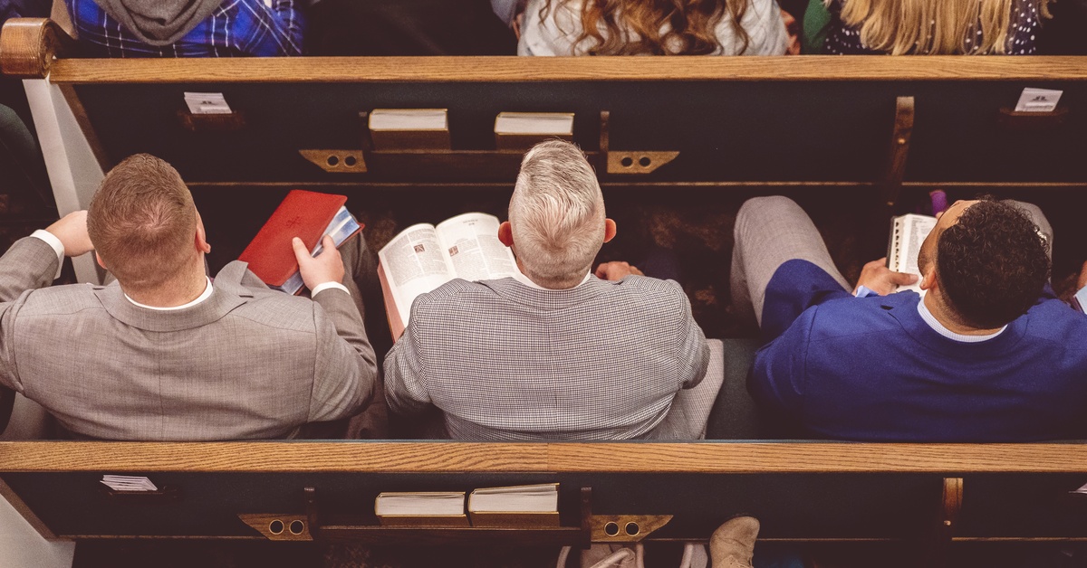Overhead view of people seated in church pews facing forward. Two men hold open Bibles, and one holds a closed Bible.