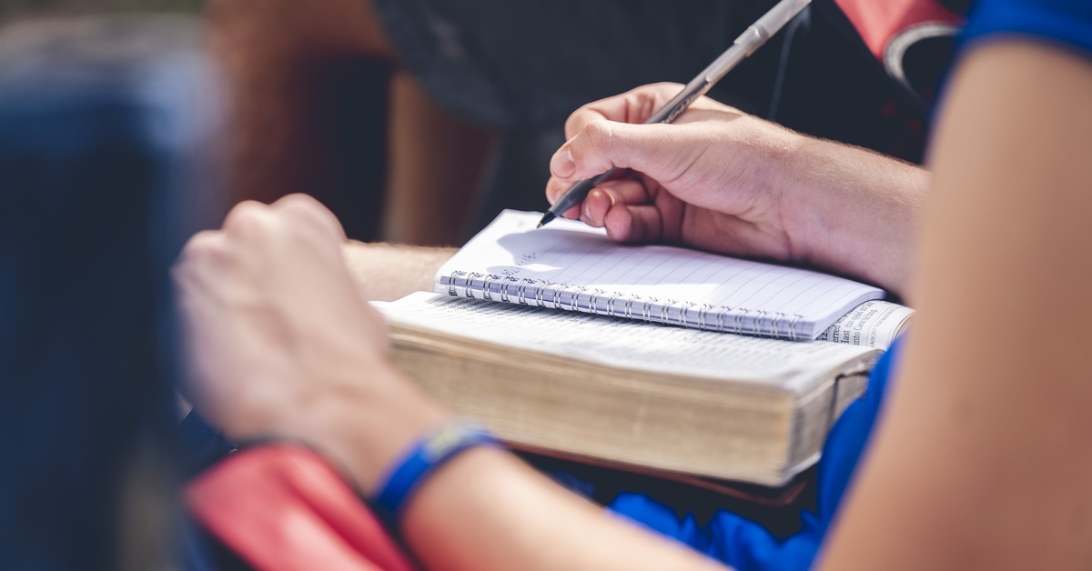 How To Get the Most out of Listening to a Sermon A close-up of a seated woman writing in a notebook, which rests on top of the open Bible in her lap.