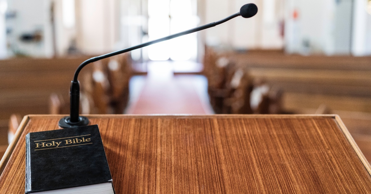 Topical vs. Expository Preaching: What’s the Difference? A hardback Bible lies closed on a wooden pulpit in front of a microphone. The wooden bench pews are empty.
