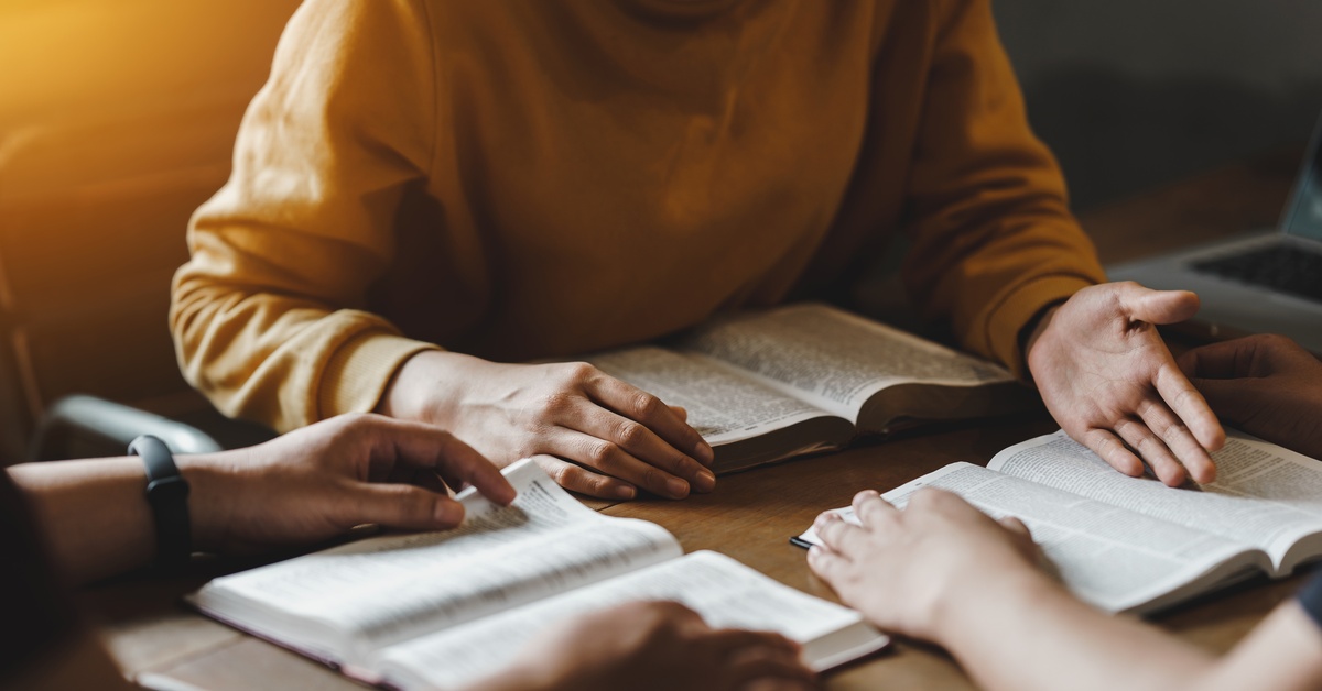 Three people sat at a table as they read the Bible together in deep discussion. One person wear a yellow sweater.