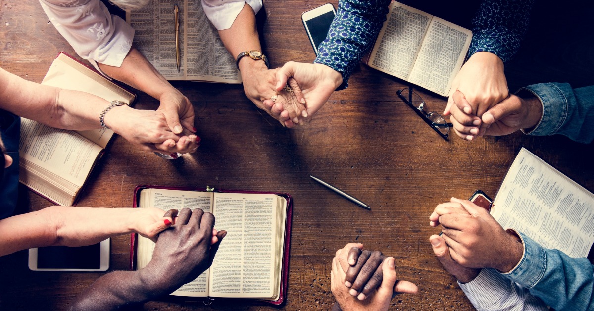 A top-down view shows a group gathered around a table with multiple open Bibles and study materials. They hold hands.