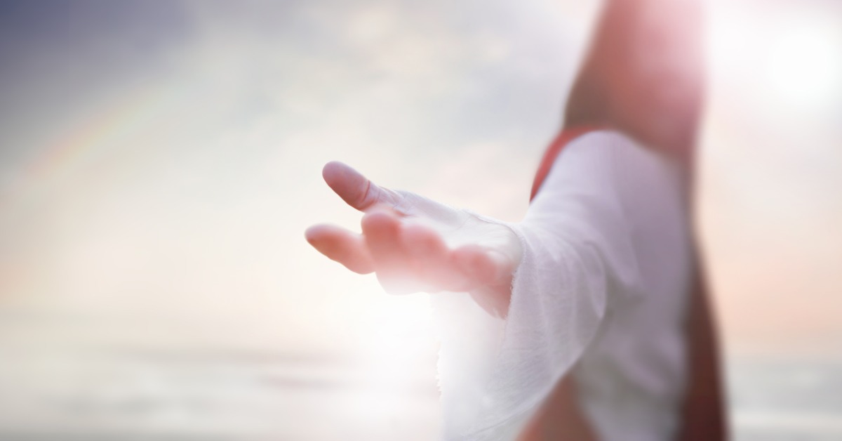 A blurred person in a white robe stretches their hand, palm-up, toward the camera. The hazy background is illuminated.
