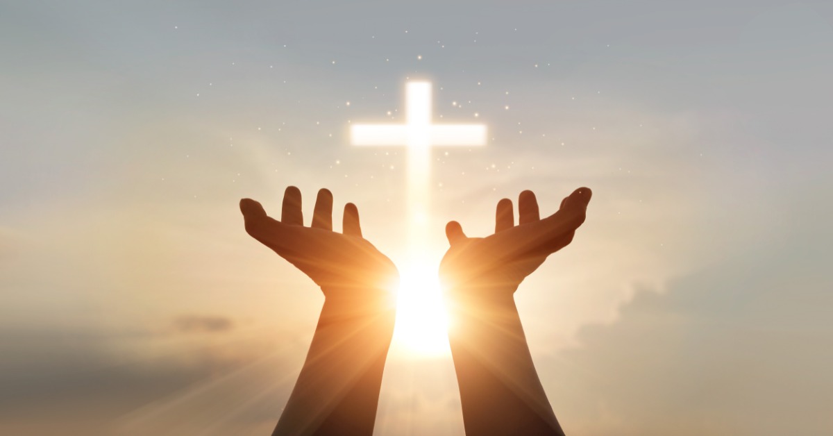 Two hands are raised against a bright sunrise, palms open toward the sky. A glowing cross appears above the hands.