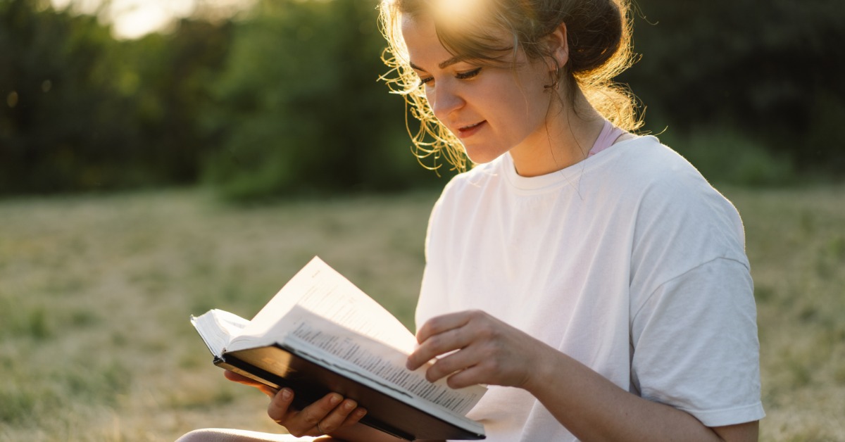 A young woman sits outdoors in a grassy field and turns a book's pages. Sunlight shines behind her, casting a warm glow.