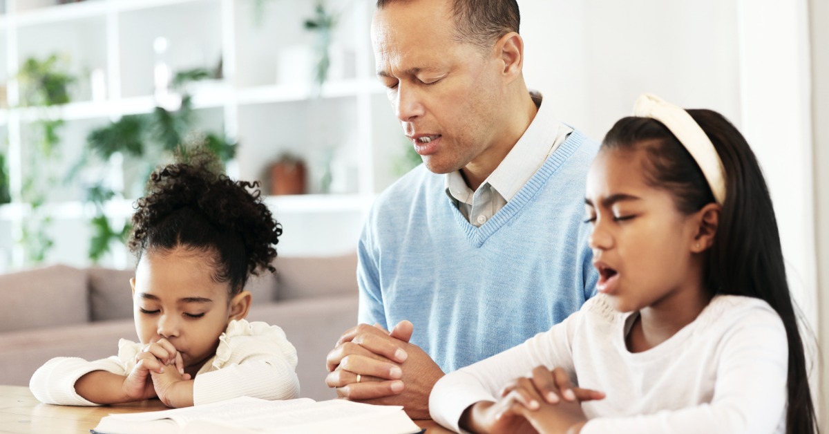 A man and two girls sit at a table with their hands clasped and eyes closed in prayer. An open book lies before them.