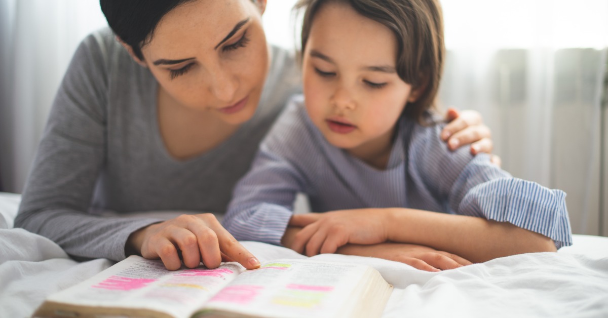 A woman gently rests her arm around a child as they read a book together. She points to a line on the highlighted page.