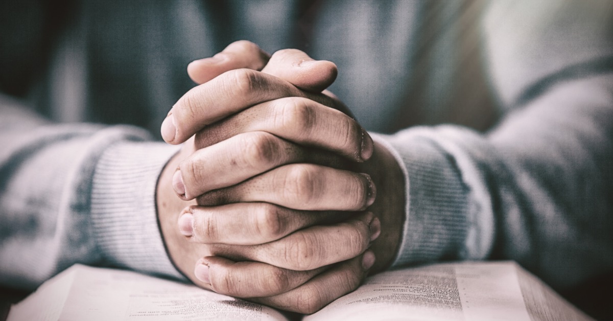 A close-up of a person&rsquo;s hands clasped together, resting on an open book. They are wearing a long-sleeve gray shirt.