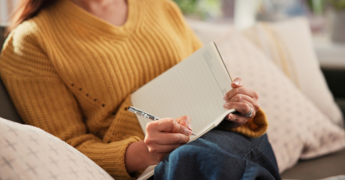 A close-up of someone writing in a blank notebook while seated. They are wearing a textured yellow sweater and jeans.