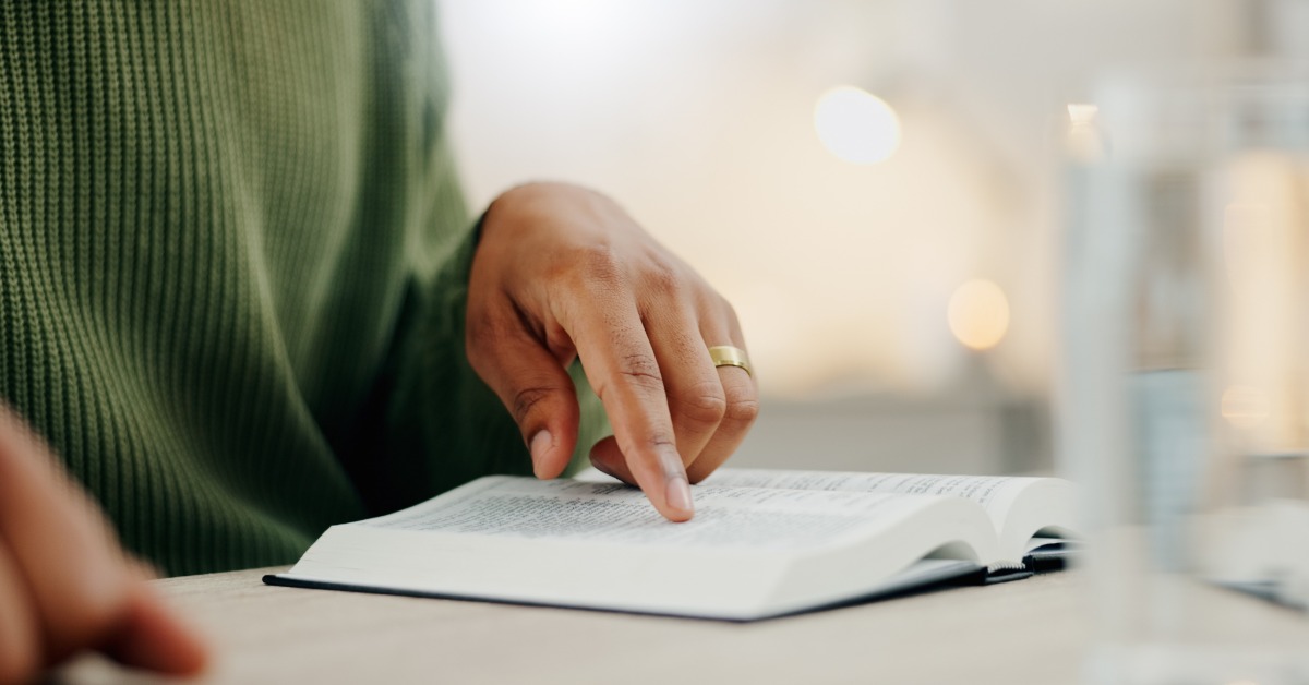 A person wearing a green sweater points to a line in an open book. The table is white, the background blurred.