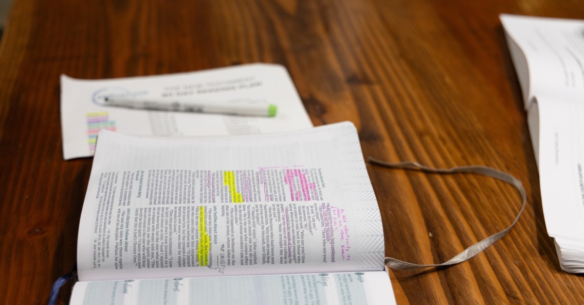 A wide-margin Bible with highlighted verses and written notes is on a wooden table. A pen and loose paper are near it.