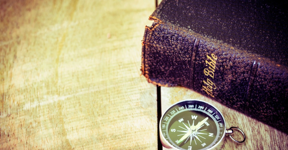 A worn Bible with a textured black cover lies on a rustic wood surface. A black-and-silver compass lies near the book.