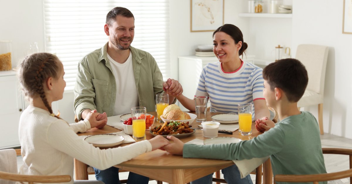 8 Ways To Worship God in Your Everyday Life Two adults and two children hold hands as they sit together at a table with food and drinks in front of them.