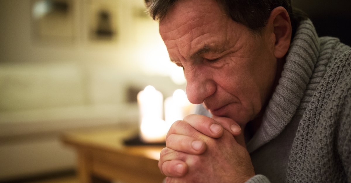 A man sits with his eyes closed, head bowed, and hands clasped under his chin. Warm electric candles light the room.