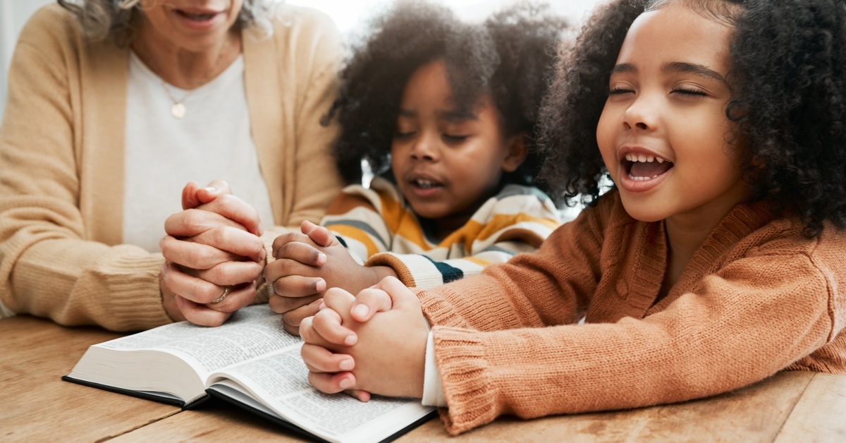 A woman and two kids sit with their eyes closed and hands clasped in prayer. An open book is in front of them. A woman and two kids sit with their eyes closed and hands clasped in prayer. An open book is in front of them.