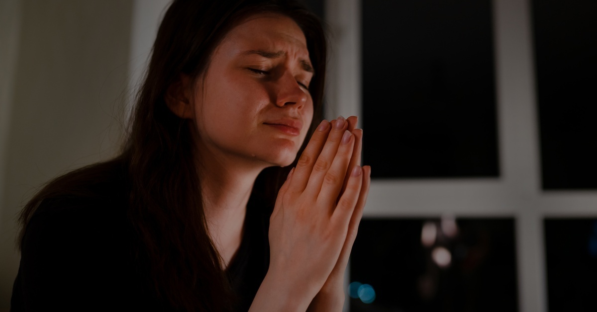 A woman sits near a dark window with her eyes closed, her brows furrowed, and her hands clasped in prayer. A woman sits near a dark window with her eyes closed, her brows furrowed, and her hands clasped in prayer.