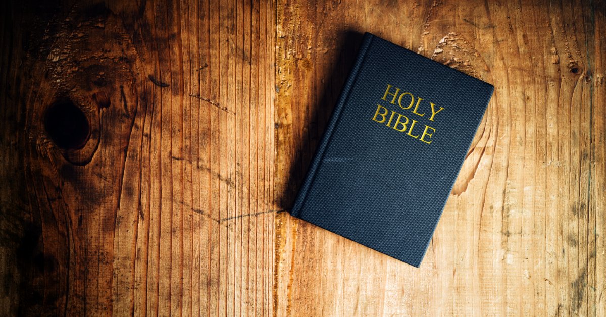 A dark-covered Bible lies closed on a weathered wood surface. The words &ldquo;Holy Bible&rdquo; are printed in gold on the cover.