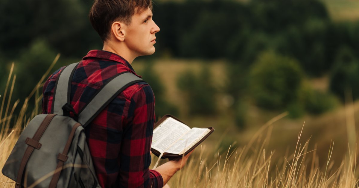 A man wearing a backpack holds open a book with dual columns printed on the pages. He stands among tall, golden grass.