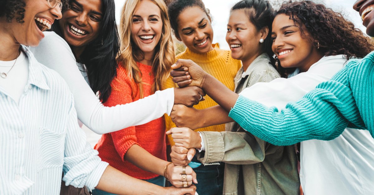 Several smiling women stand shoulder to shoulder. They each put a hand in the center of the group, one over the other.