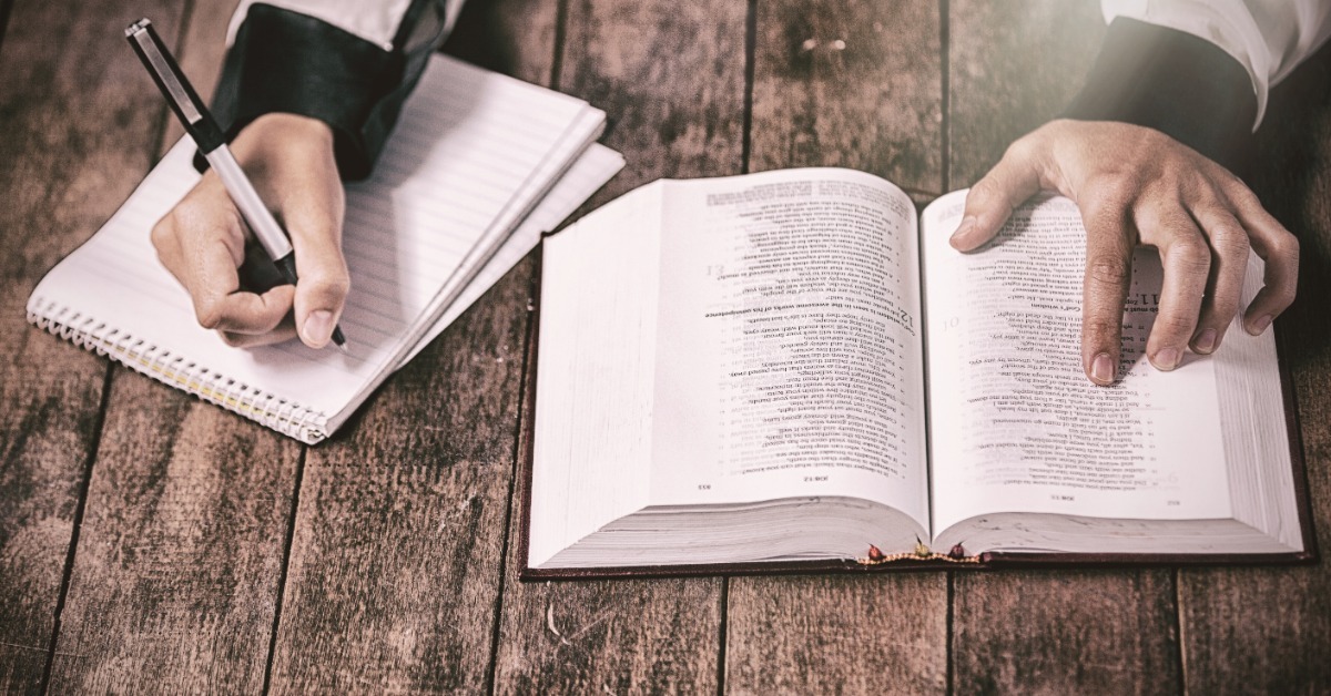 A person keeps an index finger under a Bible verse. In their other hand, they hold a pen over a spiral-bound notebook.