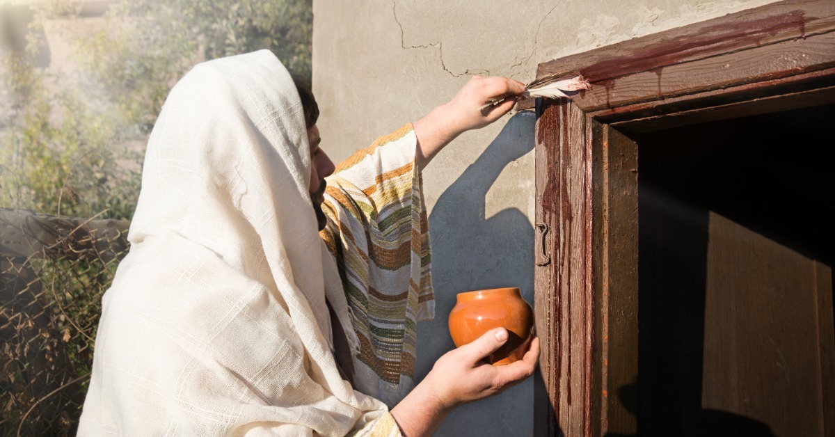 A person in a head covering holds a jar in their right hand. With their left hand, they brush liquid onto a doorframe.