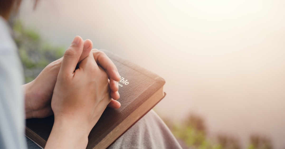 A close-up shows a person's clasped hands resting on top of a leather-bound Bible with gold lettering on the cover.
